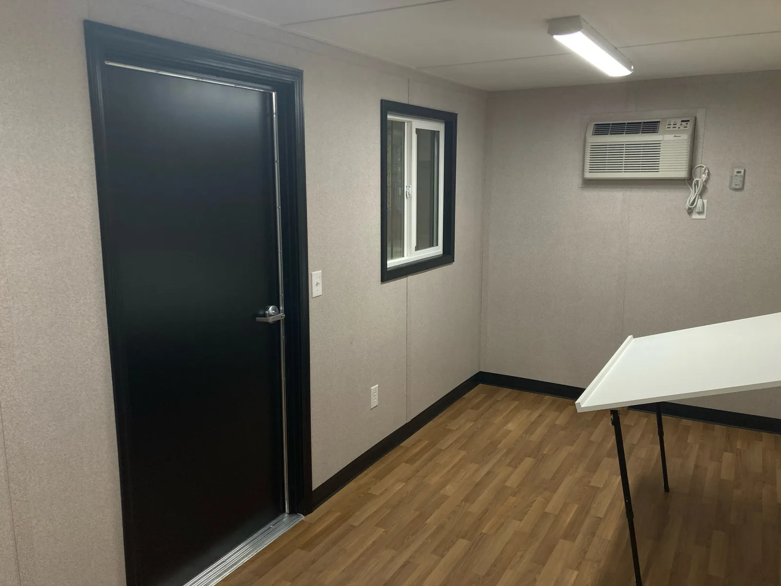 Interior of the finished office section inside a combo container — vinyl plank floor, wall-mounted AC, windows, and entry door