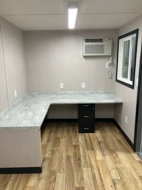 Finished charcoal container office interior with L-shaped desk, wood-look floor, window, and air conditioner
